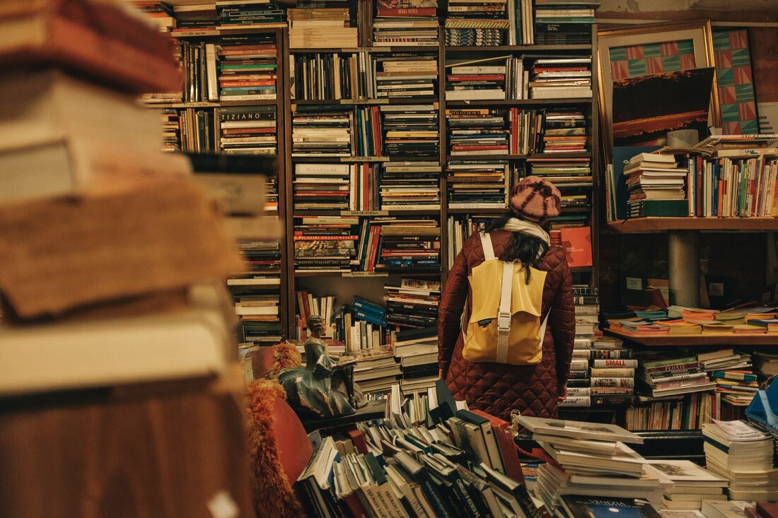 Person in front of many many haphazardly stacked books on shelves. By Darwin Vegher on Unsplash. @darwiiiin